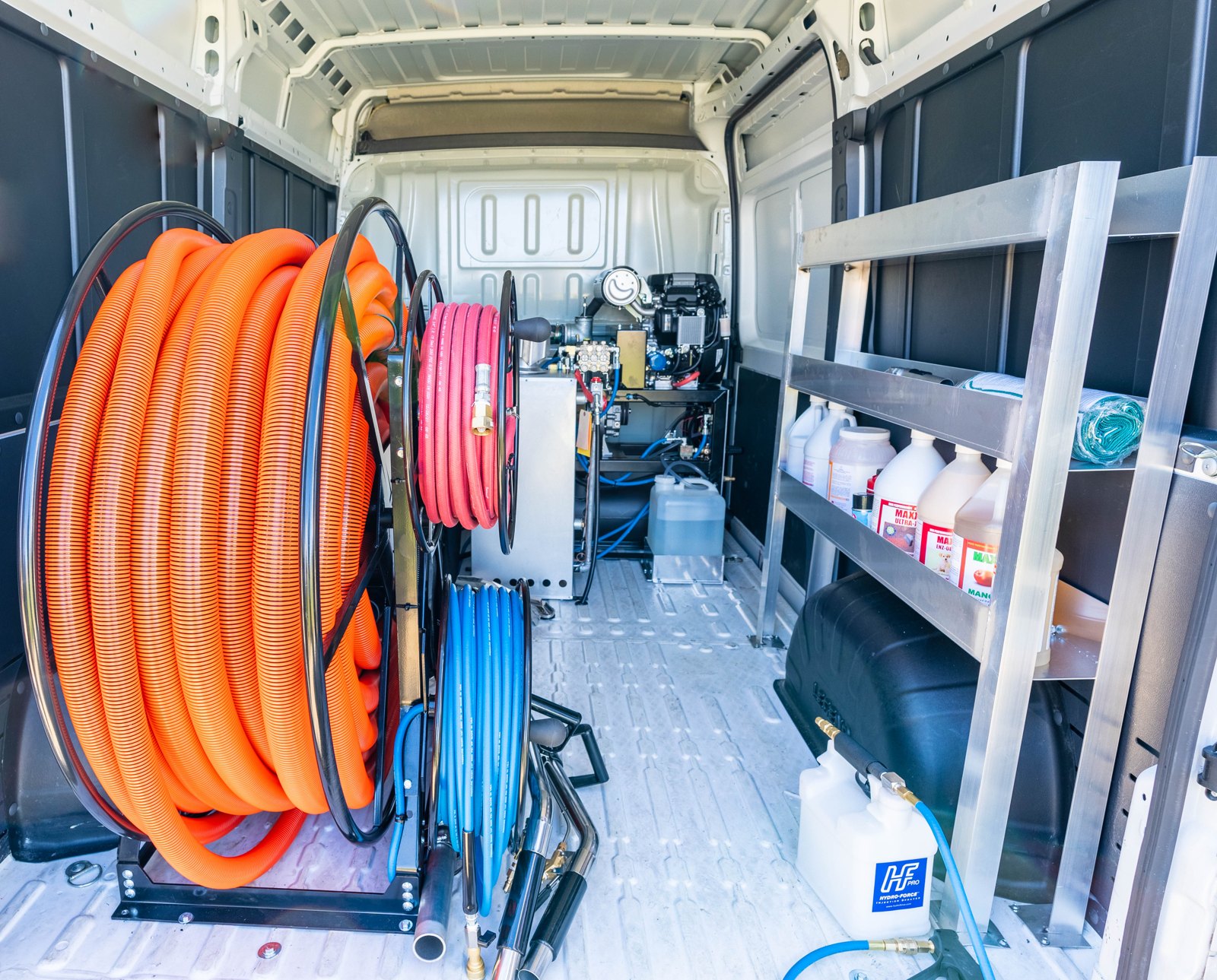 Inside the cargo van with hose reels, cleaning supplies on shelving, and the truckmount installed toward the side door.